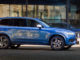Ein blauer Volvo XC90 des Modelljahres 2017 fährt an einem Bürogebäude vorbei.