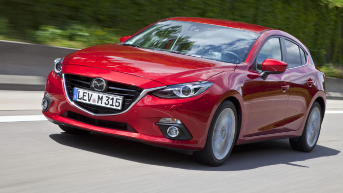 Ein roter Mazda3, fotografiert von schräg vorne, fährt auf einer Landstraße.