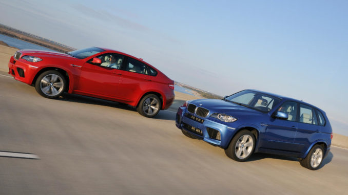 Ein roter BMW X6 M und ein blauer X5 M fahren 2009 auf einer Strandpromenade.