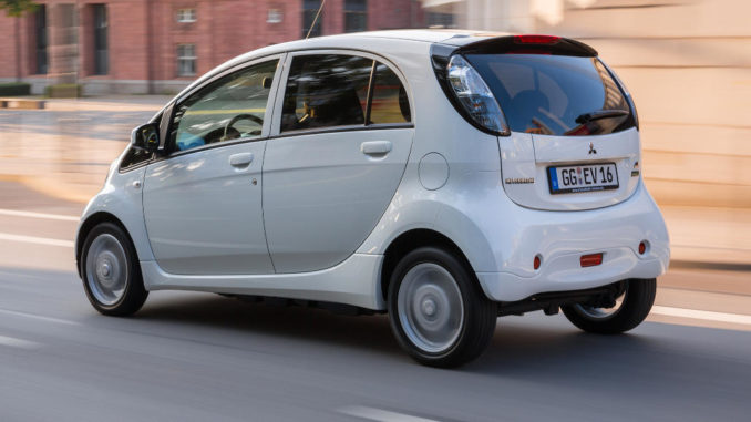 Ein weißer Mitsubishi Electric Vehicle ( I-Miev ) fährt an Backsteingebäuden vorbei.