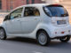 Ein weißer Mitsubishi Electric Vehicle ( I-Miev ) fährt an Backsteingebäuden vorbei.