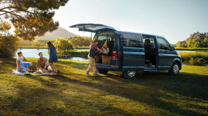 Ein blauer VW T6 Multivan steht am Rande eines Sees bei einem Familienpicknick.