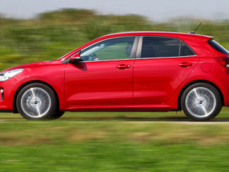 Ein roter Kia Rio fährt auf einer Landstraße an einem Feld vorbei.