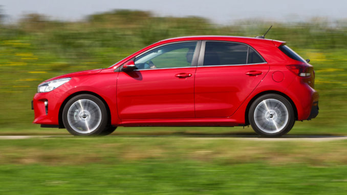 Ein roter Kia Rio fährt auf einer Landstraße an einem Feld vorbei.