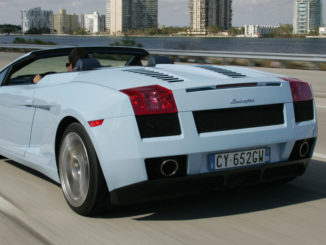 Ein hellblauer Lamborghini Gallardo Spyder fährt auf einer Uferpromenade an einigen Hochhäusern vorbei.