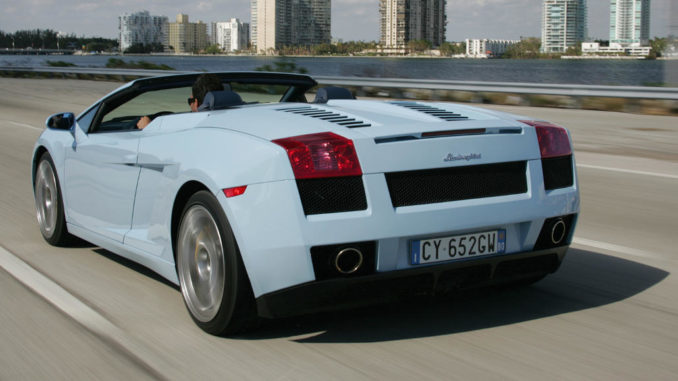 Ein hellblauer Lamborghini Gallardo Spyder fährt auf einer Uferpromenade an einigen Hochhäusern vorbei.