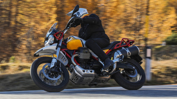 Ein Biker fährt mit einer Moto Guzzi V85 TT (2019) vor einem Herbstwald durch eine Kurve.