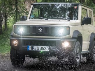 Ein beiger Suzuki Jimny fährt 2020 auf einem Waldweg durch ein Pfütze.