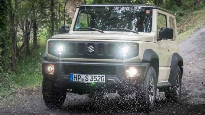 Ein beiger Suzuki Jimny fährt 2020 auf einem Waldweg durch ein Pfütze.